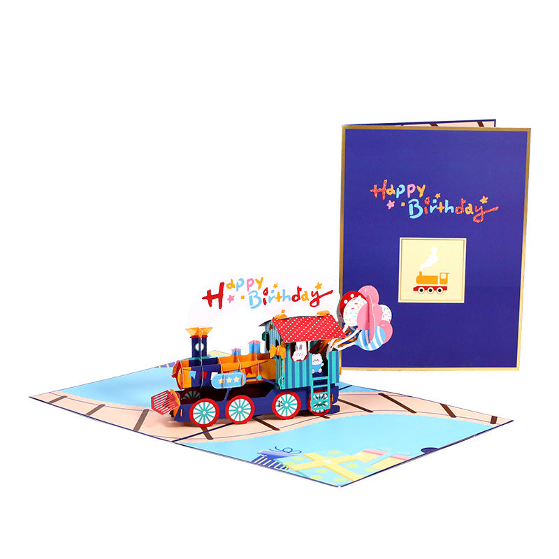 A 3D pop-up birthday card featuring a miniature train on tracks with 'Happy Birthday' text and a gift box on the cover.