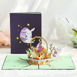 A 3D pop-up greeting card featuring a rabbit and an Easter egg with a basket of flowers, set against a dark background.