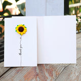 Card with a sunflower decoration and 'thank you' text on a wooden surface.