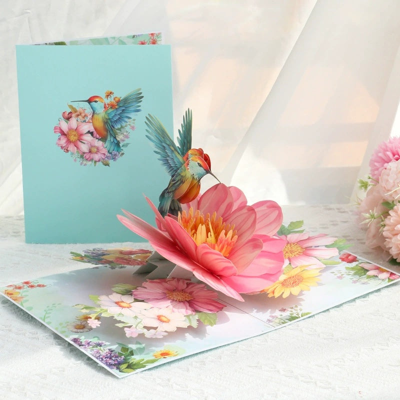 Greeting card with a pop-up design of a hummingbird and flowers on a white surface.