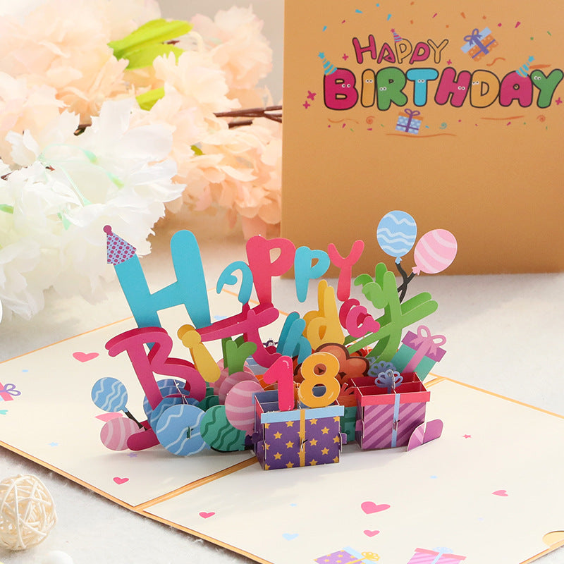 Colorful 'Happy Birthday' pop-up card with balloons and presents on a white surface.