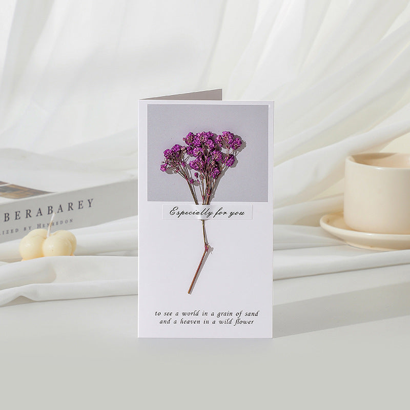 Greeting card with purple flowers on a white surface with books and a candle.