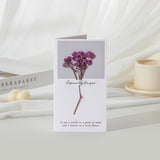 Greeting card with purple flowers on a white surface with books and a candle.