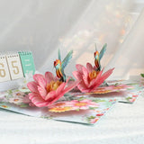 Pop-up card with pink flowers and hummingbirds on a white surface.