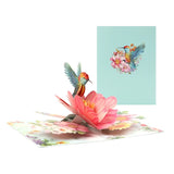 A 3D pop-up greeting card featuring a hummingbird and a flower when opened, with a printed floral pattern on the card's surface and a matching envelope.